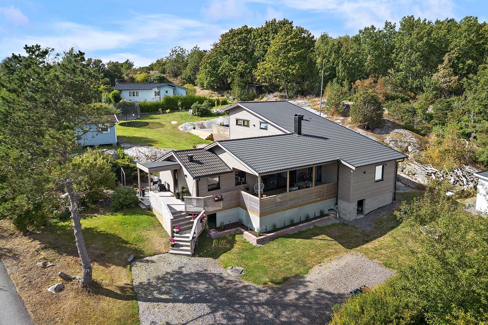 Flygbild över villa med altan och tillbyggnad, omgiven av träd och berg i dagen
